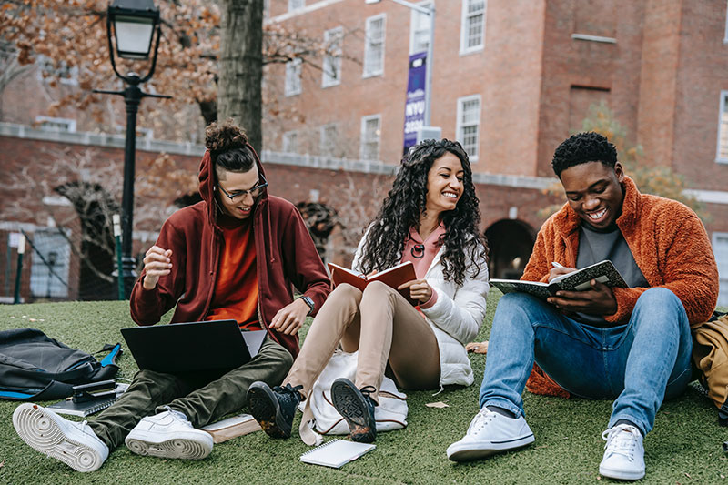 three students sitting with books on the lawn of a college campus laughing