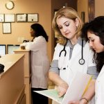 A group of four female doctors in a medical office. Two women are at the back chatting over a desk. Two women are at the front looking at a clipboard.